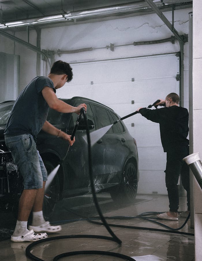 hero-img-02 Two men using hoses to clean a black car in an indoor garage.