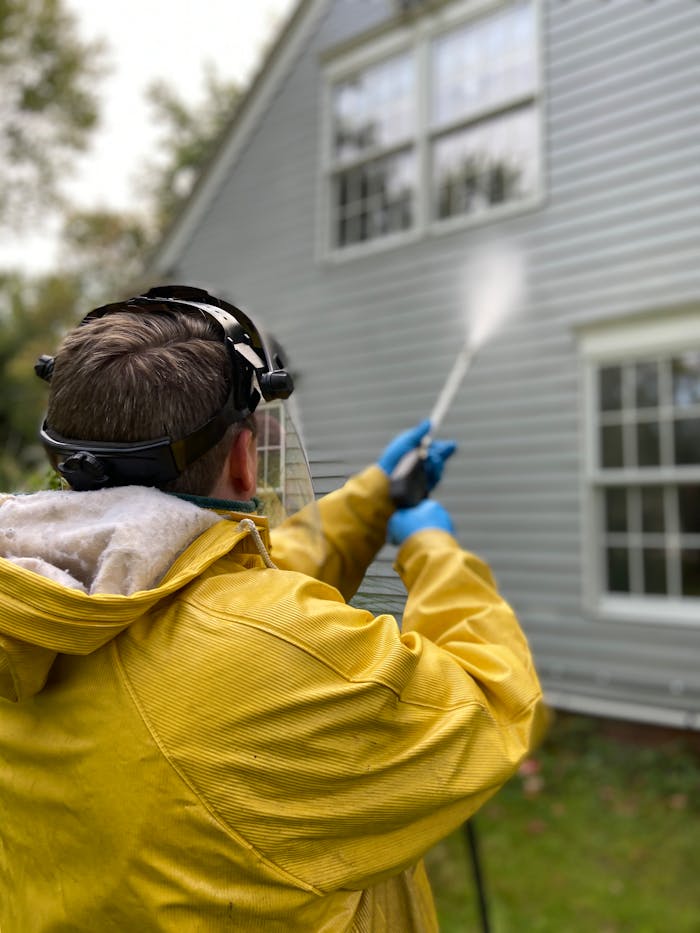 hero-img-01 Person cleaning house siding with a pressure washer, wearing protective gear in a yellow raincoat.